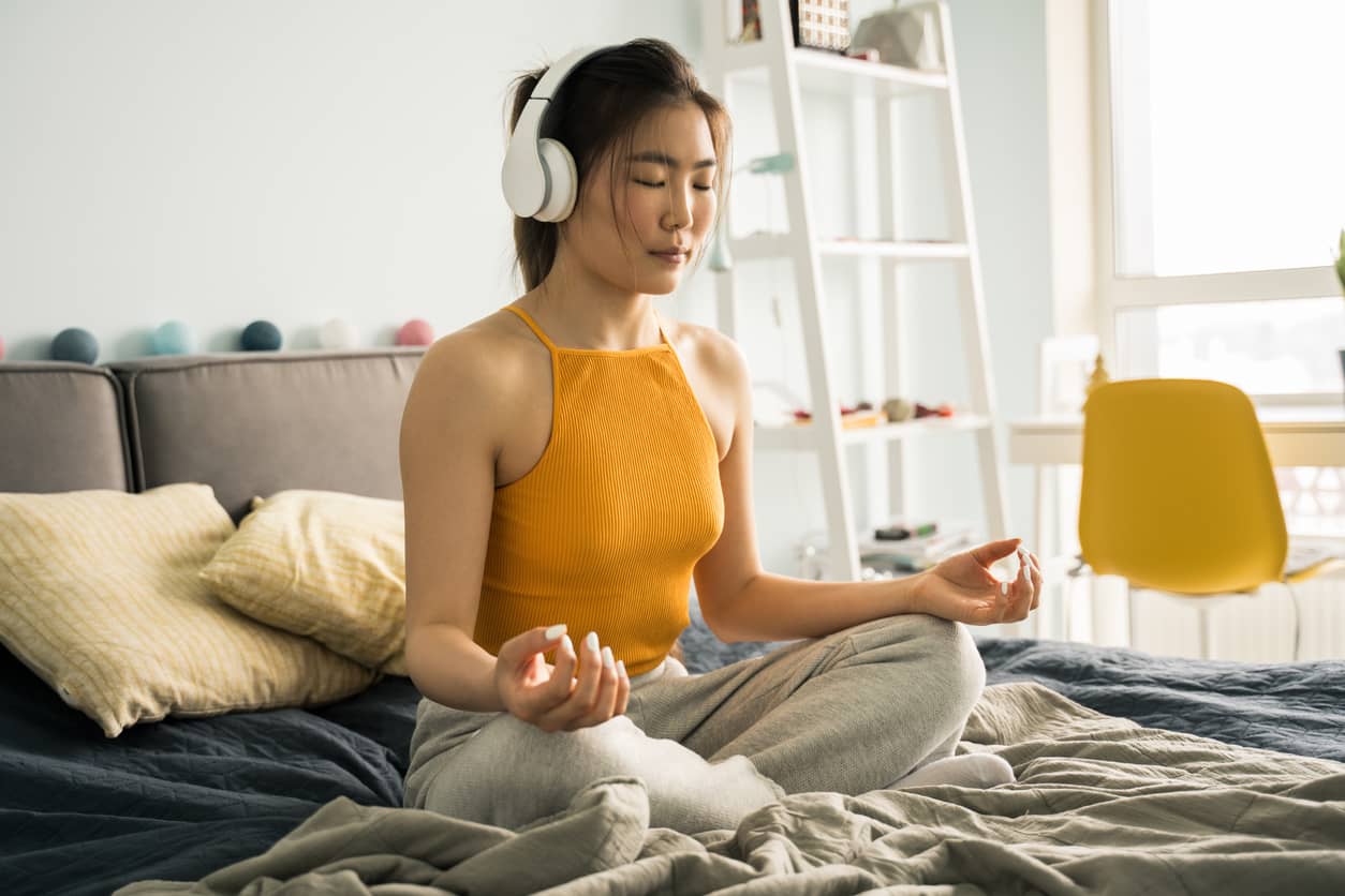 Relaxed young calm woman wearing headphones sitting at home in the bed in lotus position, while meditating and practicing yoga with pleasure smile and closed eyes. Enjoying of the morning concept